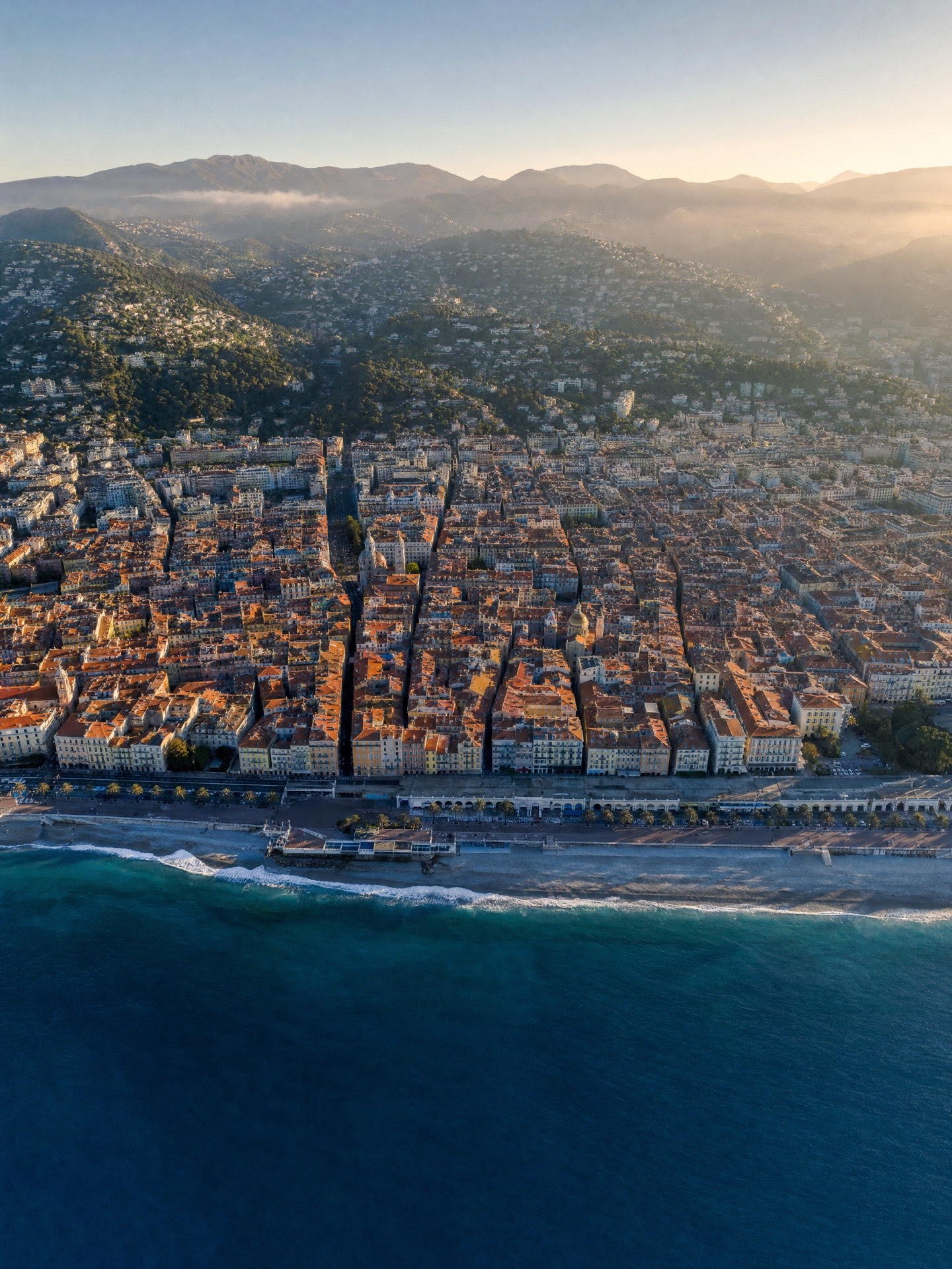 Vue aérienne de Nice au matin : la ville dense aux toits de tuiles, serrée entre la Méditerranée et les collines.